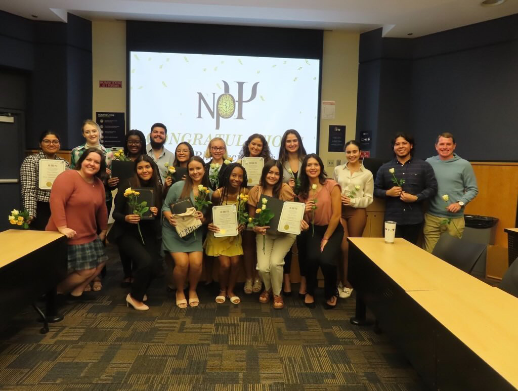 Florida International University’s induction ceremony. There are three rows of members, most of whom are holding white roses. The new inductees are holding folders that contain their induction certificates. They are smiling in the background is a blue wall and projector screen on that wall that has a white background, the Nu Rho Psi insignia, confetti coming from the top, and it appears to read “congratulations”. (Some of the members are blocking the screen’s readability).