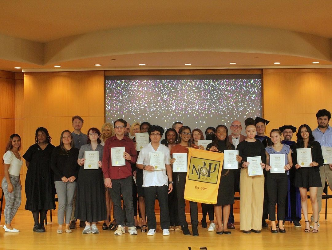 Georgia State University Nu Rho Psi students and faculty lined up on auditorium stage. The auditorium is a golden wood color on the floor and the background and the ceiling. There is a projector screen behind everybody that has a black background and confetti, falling down from the top. The front row of students are holding their Nu Rho Psi induction certificates. The two center most students are also holding the Nu Rho Psi banner. In the back of the group are the faculty members of the GSU Nu Rho Psi chapter.