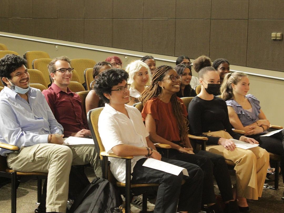 Students/new membres during the Georgia State University fall 2023 Nu Rho Psi induction ceremony. There are three rows of students sitting in golden colored seats in the auditorium that the induction ceremony is held in. This photo was taken likely during the ceremony of the students and they are all looking towards the stage.