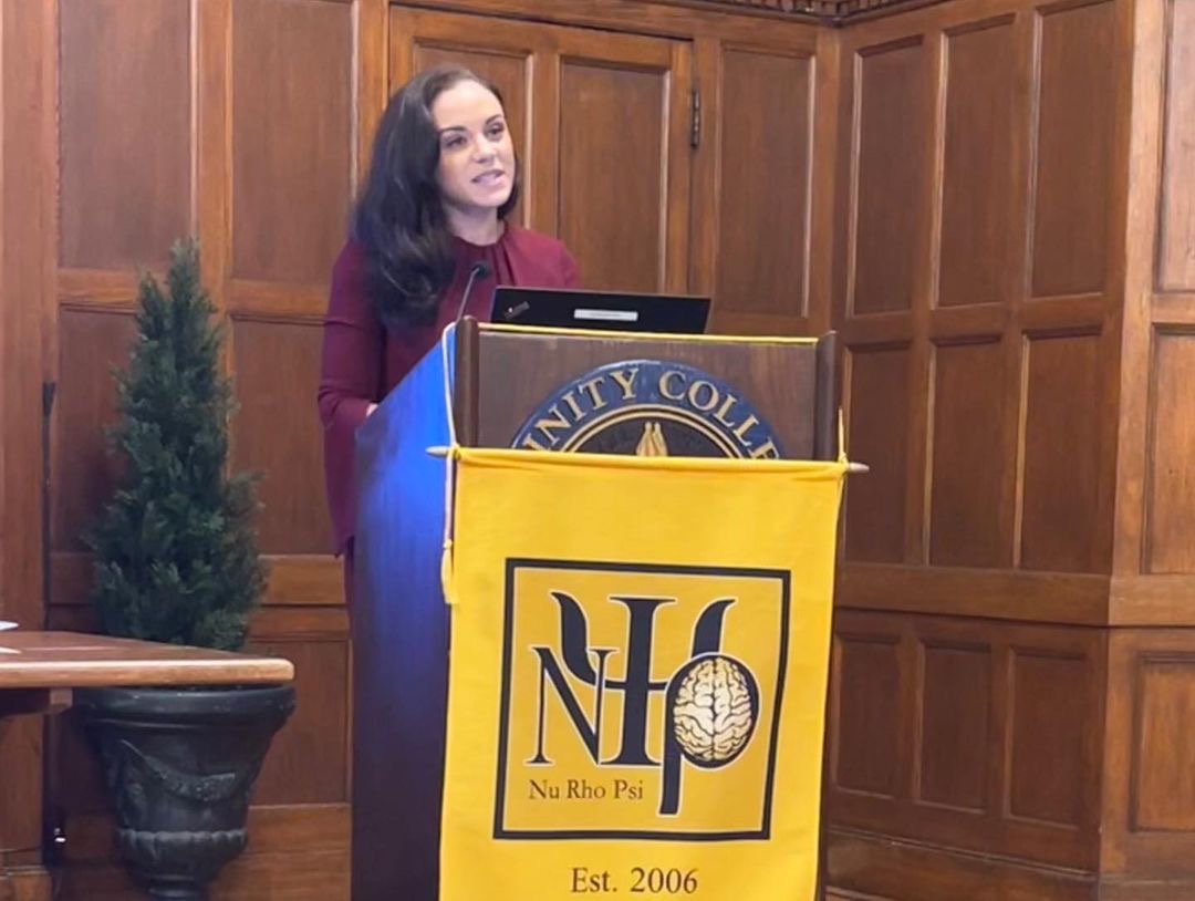 Induction from the Trinity College Nu Rho Psi chapter during the ceremony of Dr. Ginger Mills delivering the keynote. Dr. Ginger Mills is standing behind a podium in the middle of speaking. The podium has the Trinity college insignia on the front of it, which is obscured mostly by the new banner that is hanging over top. There is a plant in the background that is similar to a small evergreen tree.