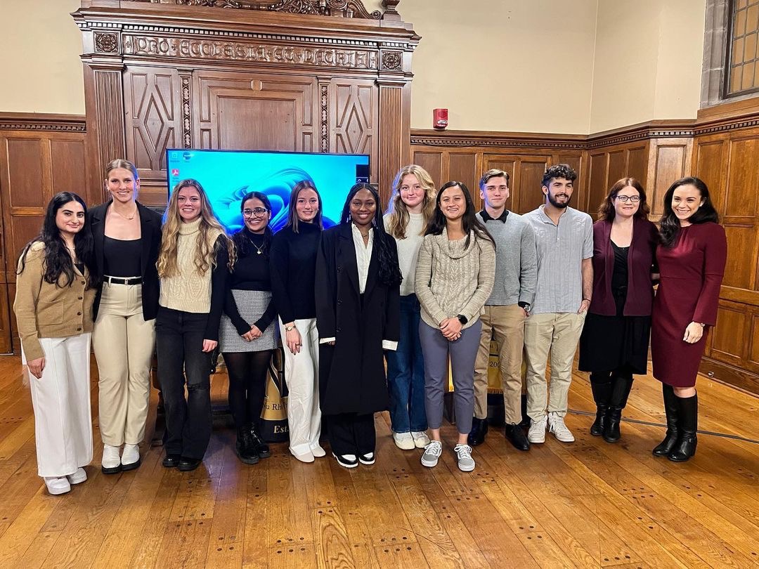 Induction from the Trinity College Nu Rho Psi chapter. There are 12 members standing together in a row and smiling - 10 students and two faculty (faculty on the rightmost side), including their keynote speaker & newly inducted honorary member, Dr. Ginger Mills.