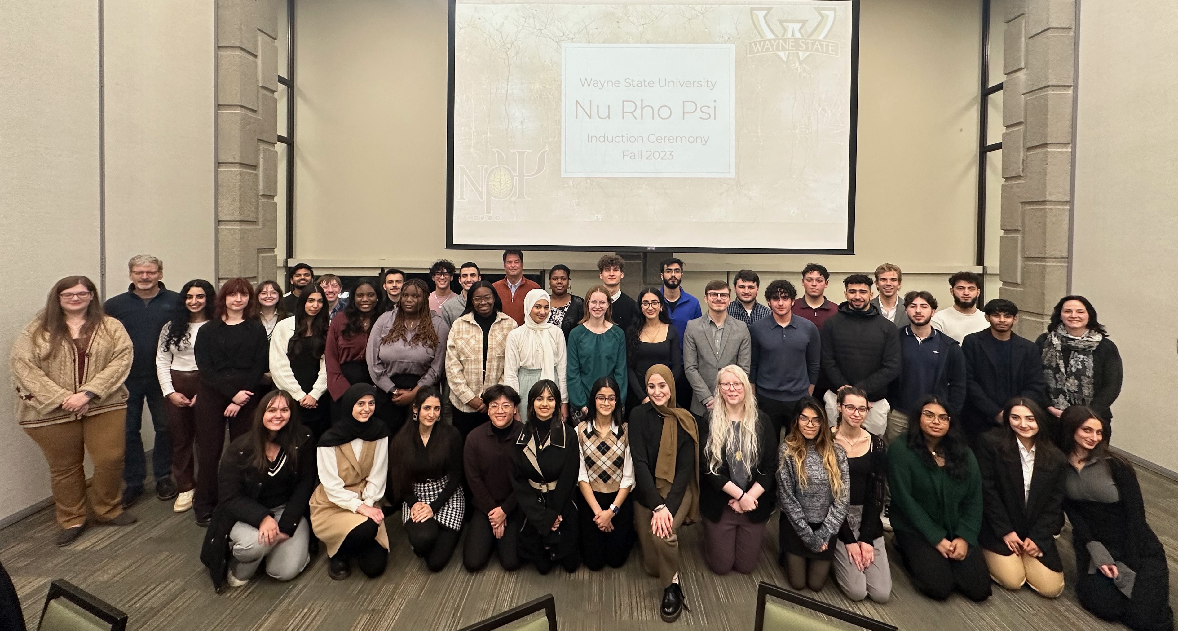 Induction from Wayne State University with the whole chapter. Above them is a slide that reads “Wayne State Nu Rho Psi Induction Ceremony Fall 2023”. There are three distinct rows of people the existing members filled in around the new members. The front row is all kneeling, while the back three rows are standing.