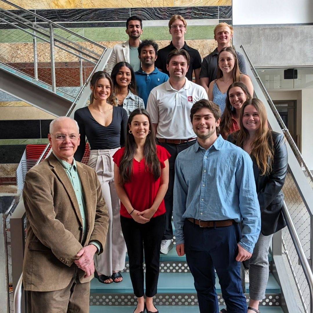 Indiana University - Bloomington’s outgoing and incoming executive board members, and the chapter advisor, at their Spring 2024 Induction ceremony. There are roughly 6 rows of students, standing on a stairwell in front of a marbled wall. They are all smiling.