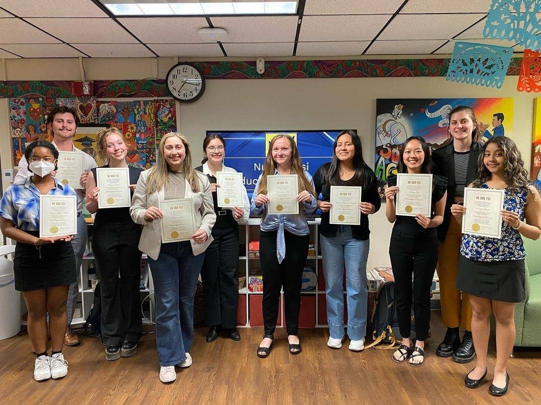 University of Arizona spring 2024 newly inducted member class. There are 10 members in a line 9 of whom are holding their induction certificates in front of them. behind the students on the left side on the wall are collages of artwork, and on the right side is a painted canvas. In the center behind the students is a screen that appears to have a slide that says Nu Rho Psi induction ceremony with the Nu Rho Psi insignia at the top.