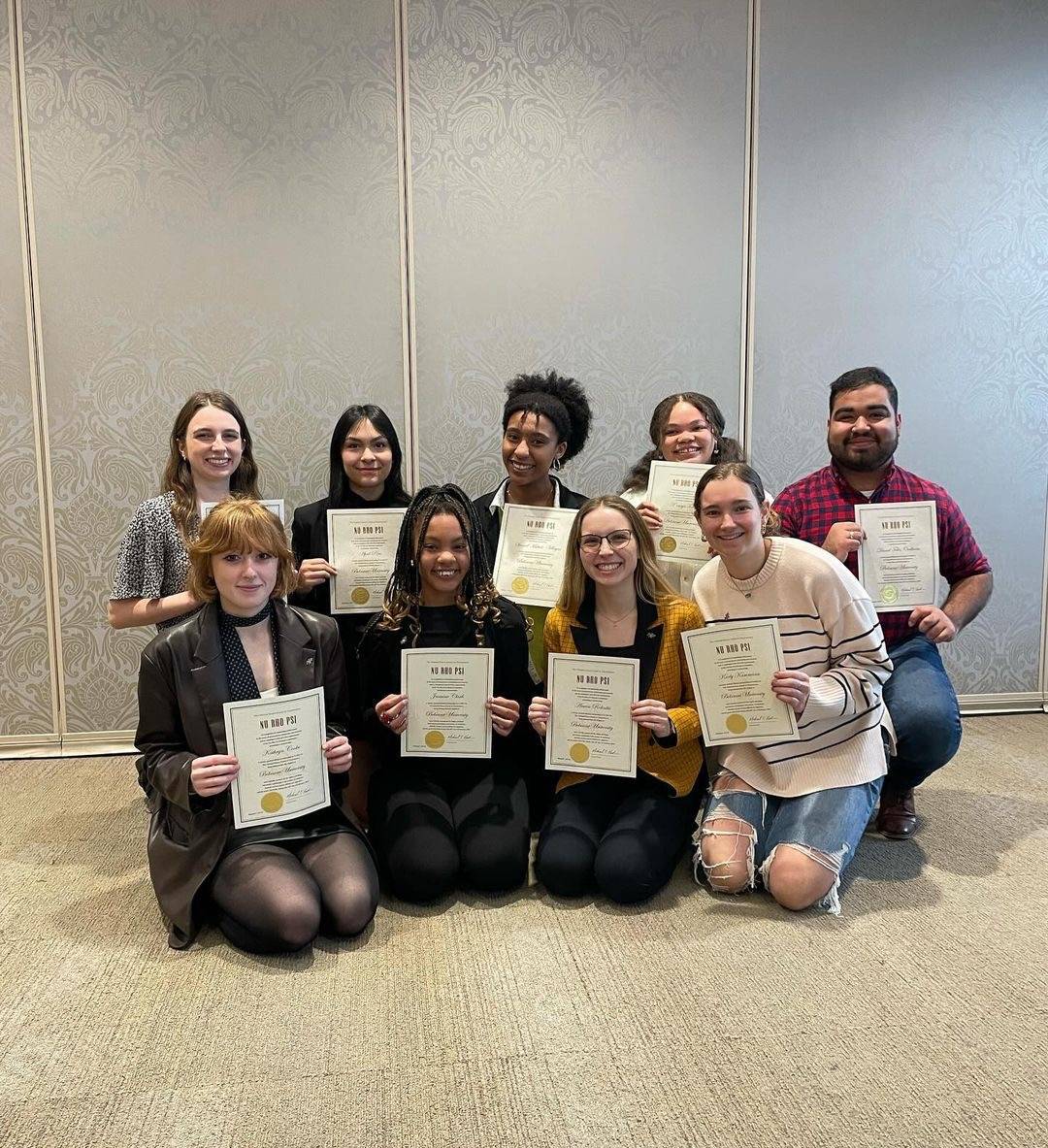 A photo of the new Belmont University, Nu Rho Psi inductees. There are nine students in total sitting in two different row. The room that they’re in has a gray wall with an ornate pattern on it and a gray beige carpet. All the students in the photo are smiling and holding their inductions certificates.