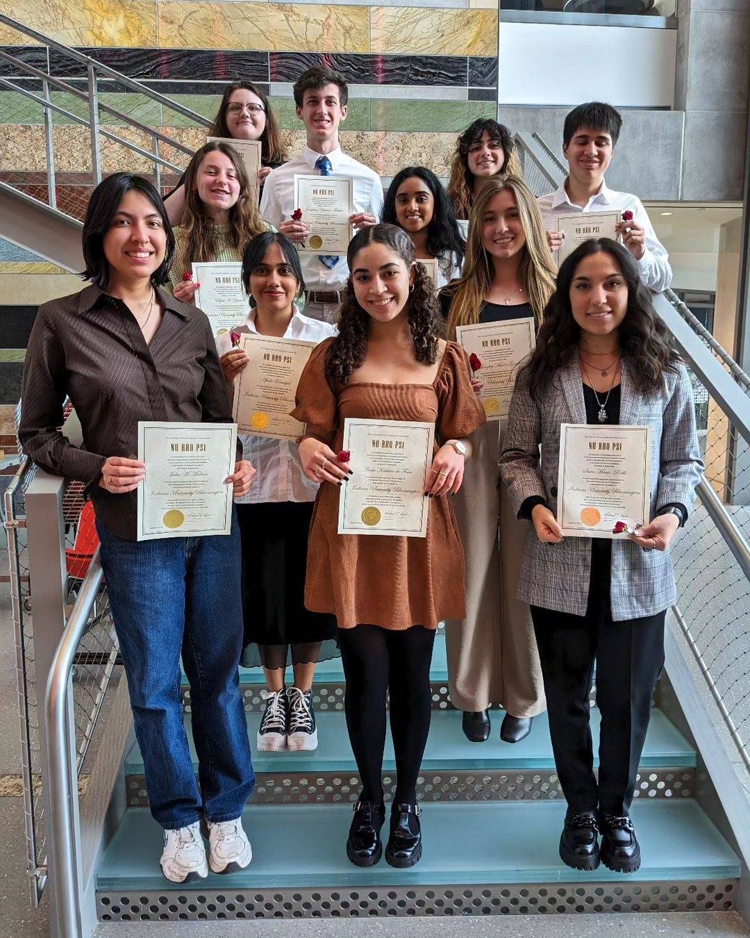 The Indiana University spring 2024 Nu Rho Psi induction ceremony a photo of the chapter’s 12 new members. The students are standing in 4 rows on a stairwell that has blue steps and silver/steel railings. The students are all smiling and holding their induction certificates and a small red rose. The new inductees featured in this photo (not in order) are: Langley Good, Isabella Flick, Anna de Four, Sara Mario Rallo, Dylan Draves, Irene Choi, Apala Poudyal, Aditi Alati, Olivia Drew, Ella Sodunke, Jonathan Matten, Julia Molina, Mahika Shetty, Ishna Kesti, Elyssa Hyman, Hailey Harper, and Adriana Lara