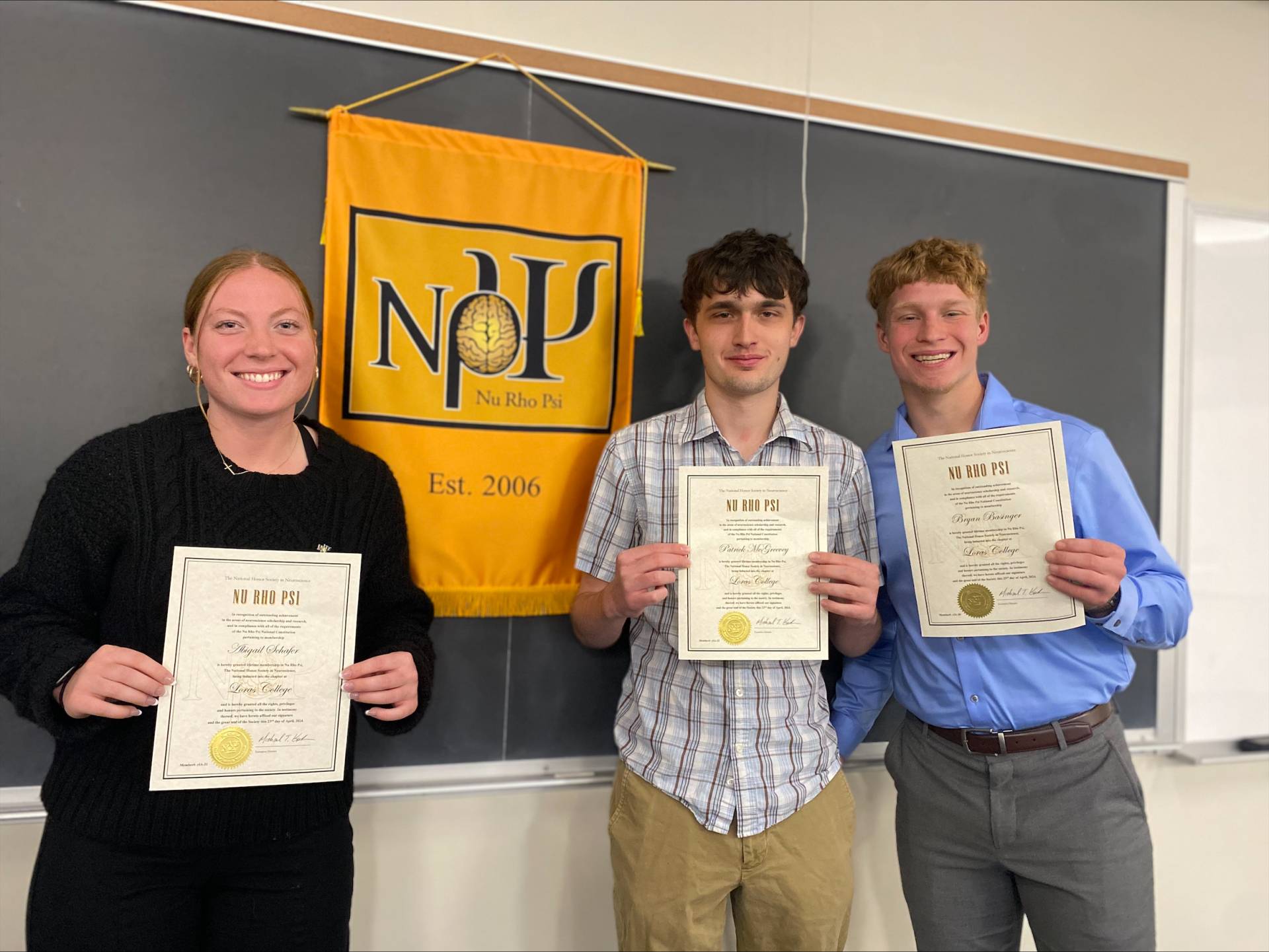 Loras College’s three newest Spring 2024 inductees: Abigail Schafer, Patrick McGreevy, and Bryan Basinger, standing in front of a chalkboard that has the Nu Rho Psi golden banner hanging up. They are holding their certificates and smiling